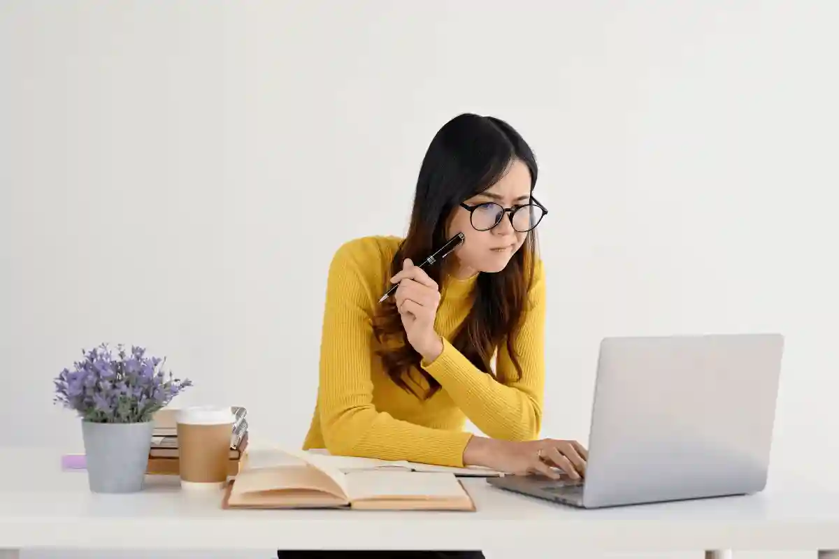 seorang wanita berkemeja kuning dan berkacamata duduk di depan meja dengan laptop